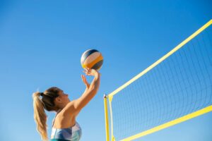 A woman plays volleyball on a sunny day, reaching to hit the ball over the net.